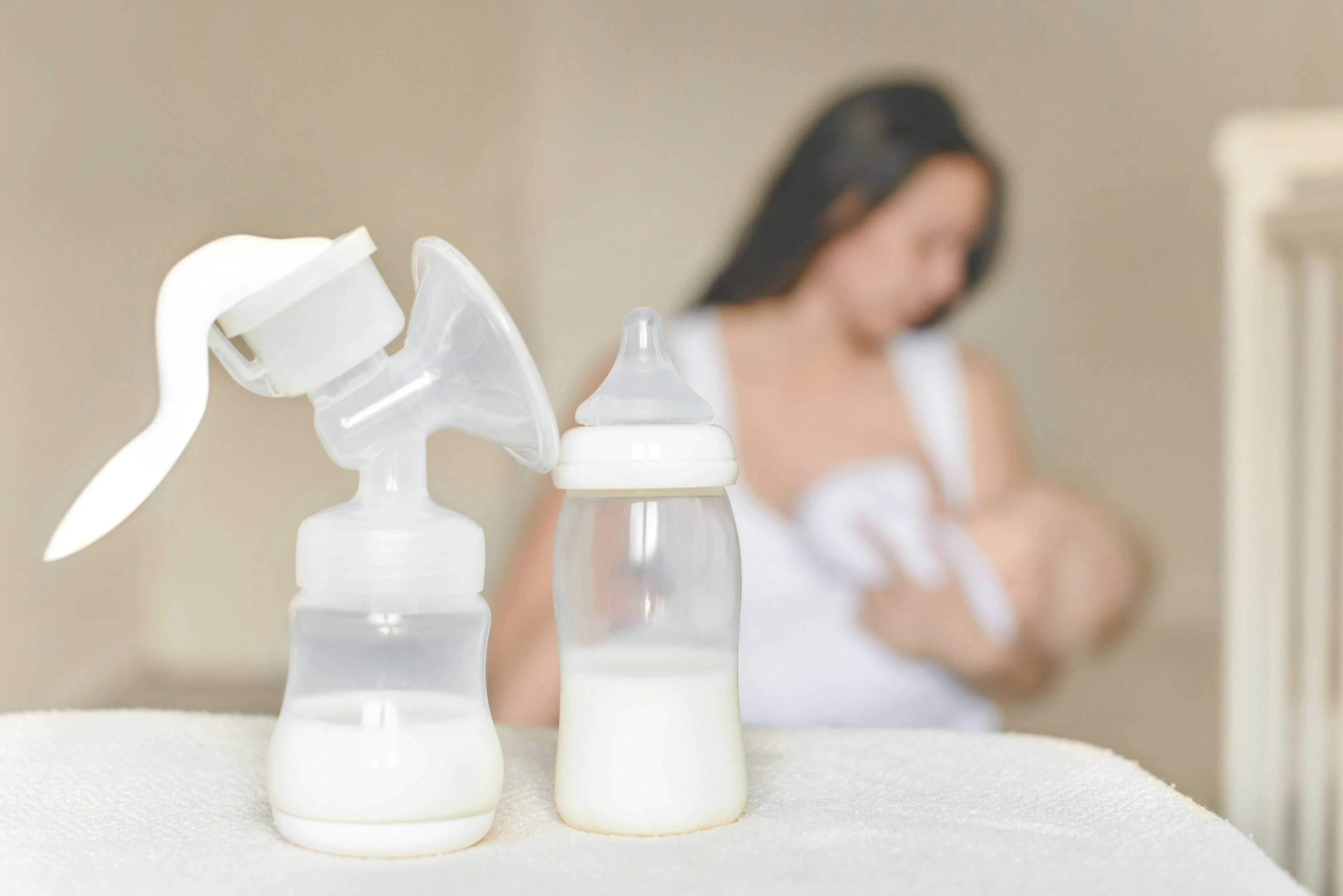 Manual breast pump and bottle with breast milk on the background of mother holding in her hands and breastfeeding baby. Maternity and baby care - Image credit: evso | stock.adobe.com