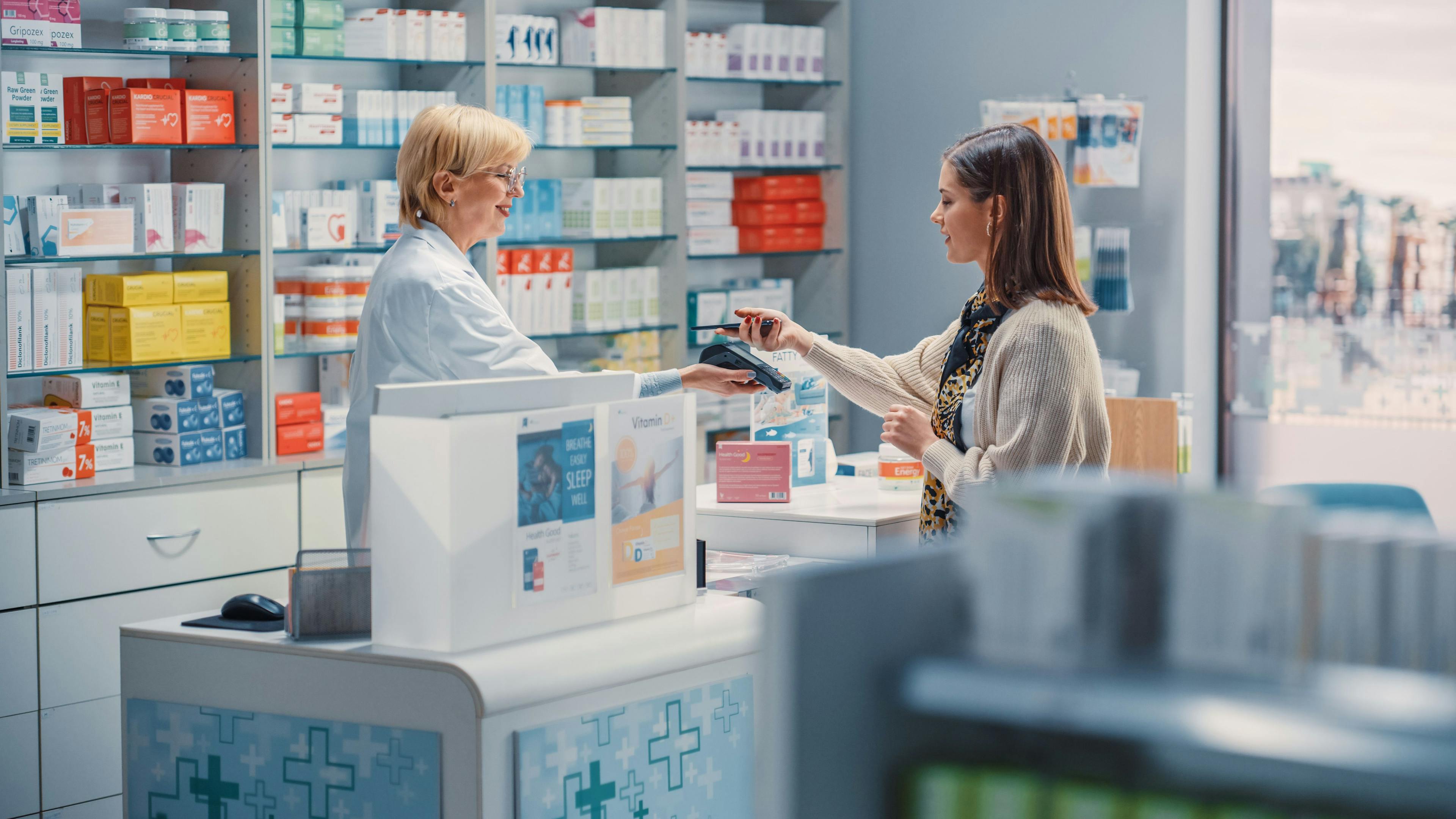 Pharmacy Drugstore Checkout Cashier Counter: Mature Female Pharmacist and Young Woman Using Contactless Payment NFC Smartphone to Buy Prescription Medicine, Vitamins, Beauty, Health Care Products - Image credit: Gorodenkoff | stock.adobe.com