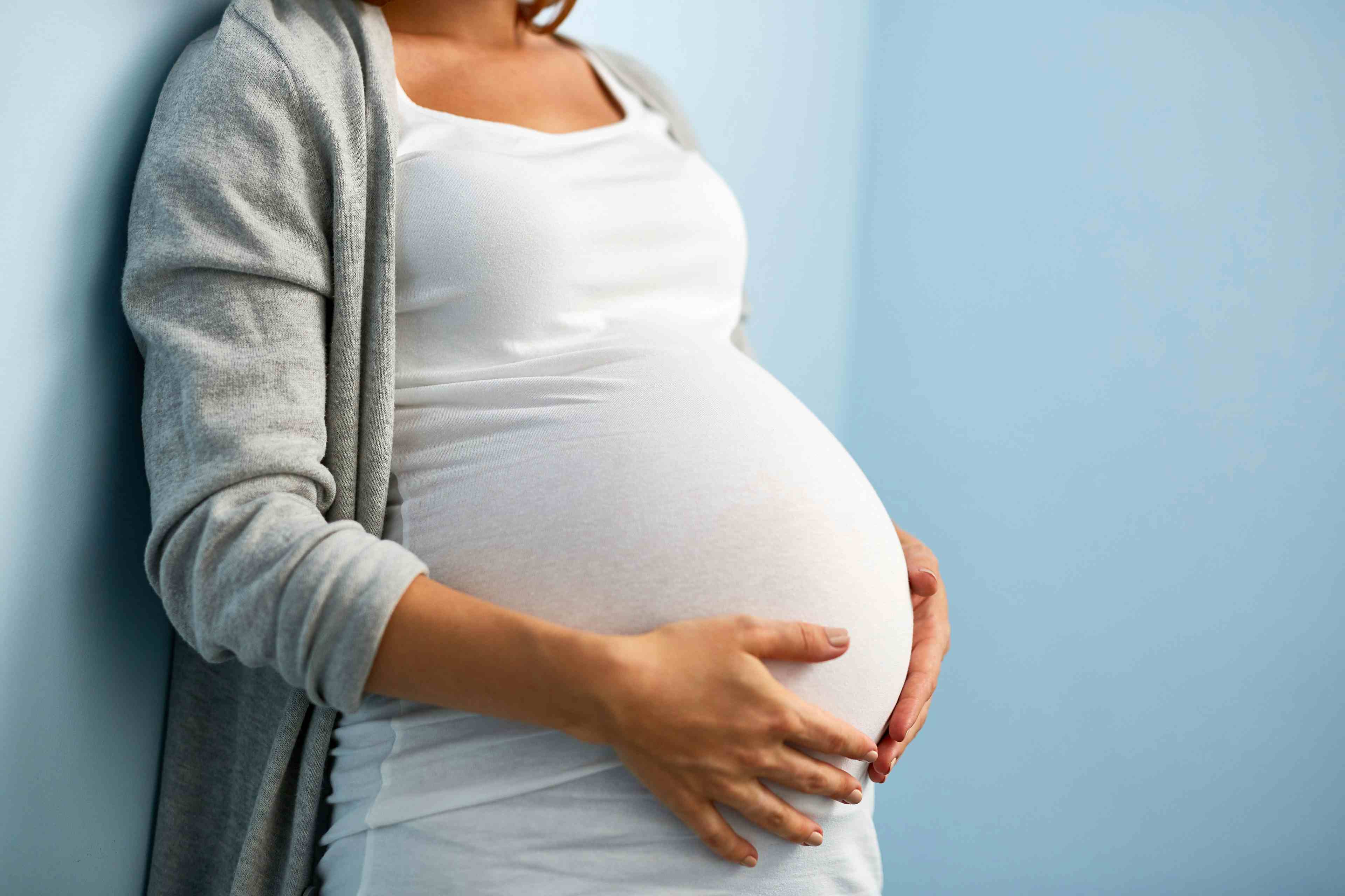 Mid-section portrait of unrecognizable woman during last months of pregnancy holding her big belly gently standing against wall in blue room - Image credit: pressmaster | stock.adobe.com
