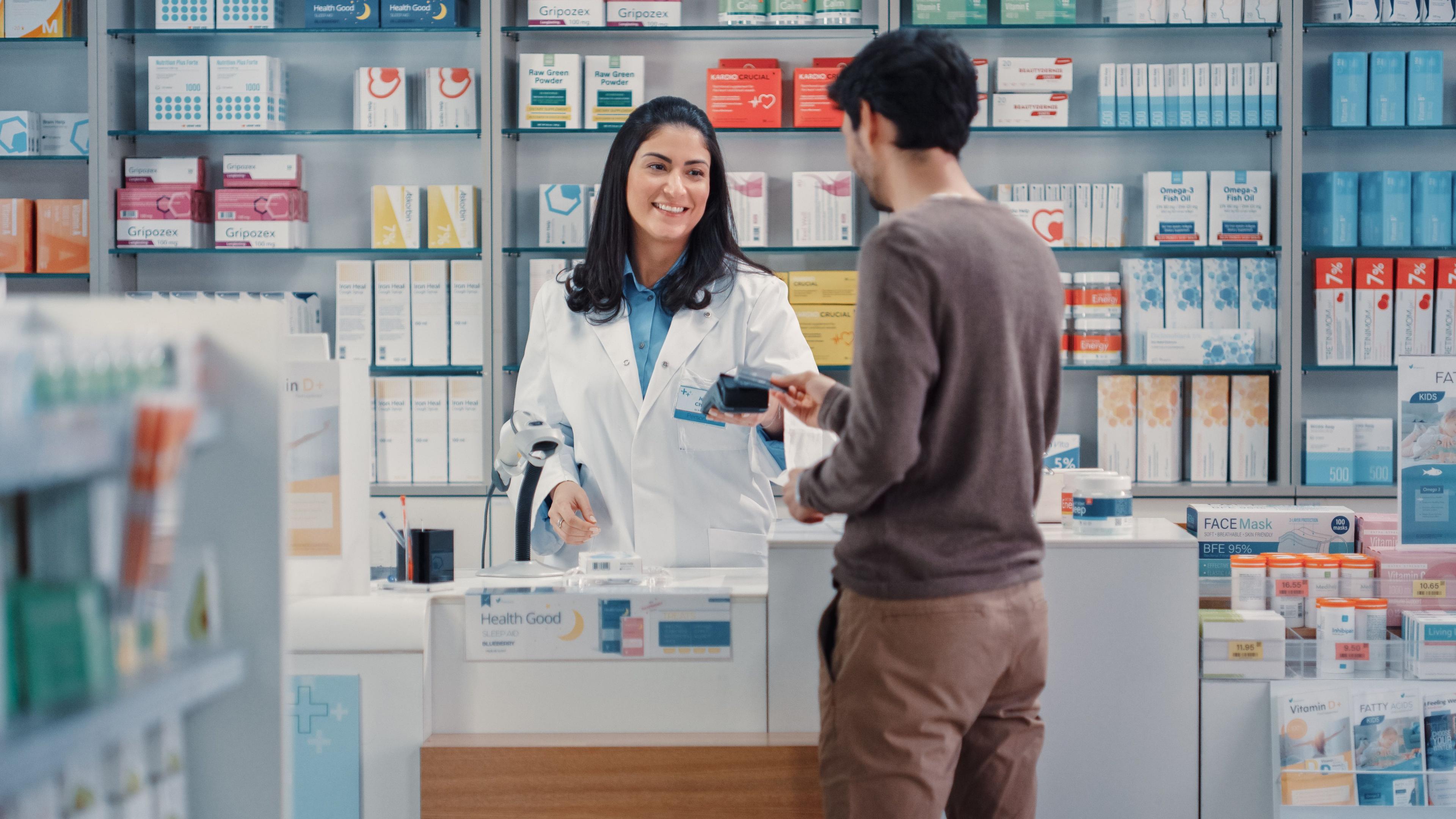 Pharmacy Drugstore: Man Chooses Medicine, Comes to the Counter, Talks to Beautiful Pharmacist Cashier, Pays for the Health Care Products at the Checkout Counter Using Contactless Payment Credit Card - Image credit: Gorodenkoff | stock.adobe.com