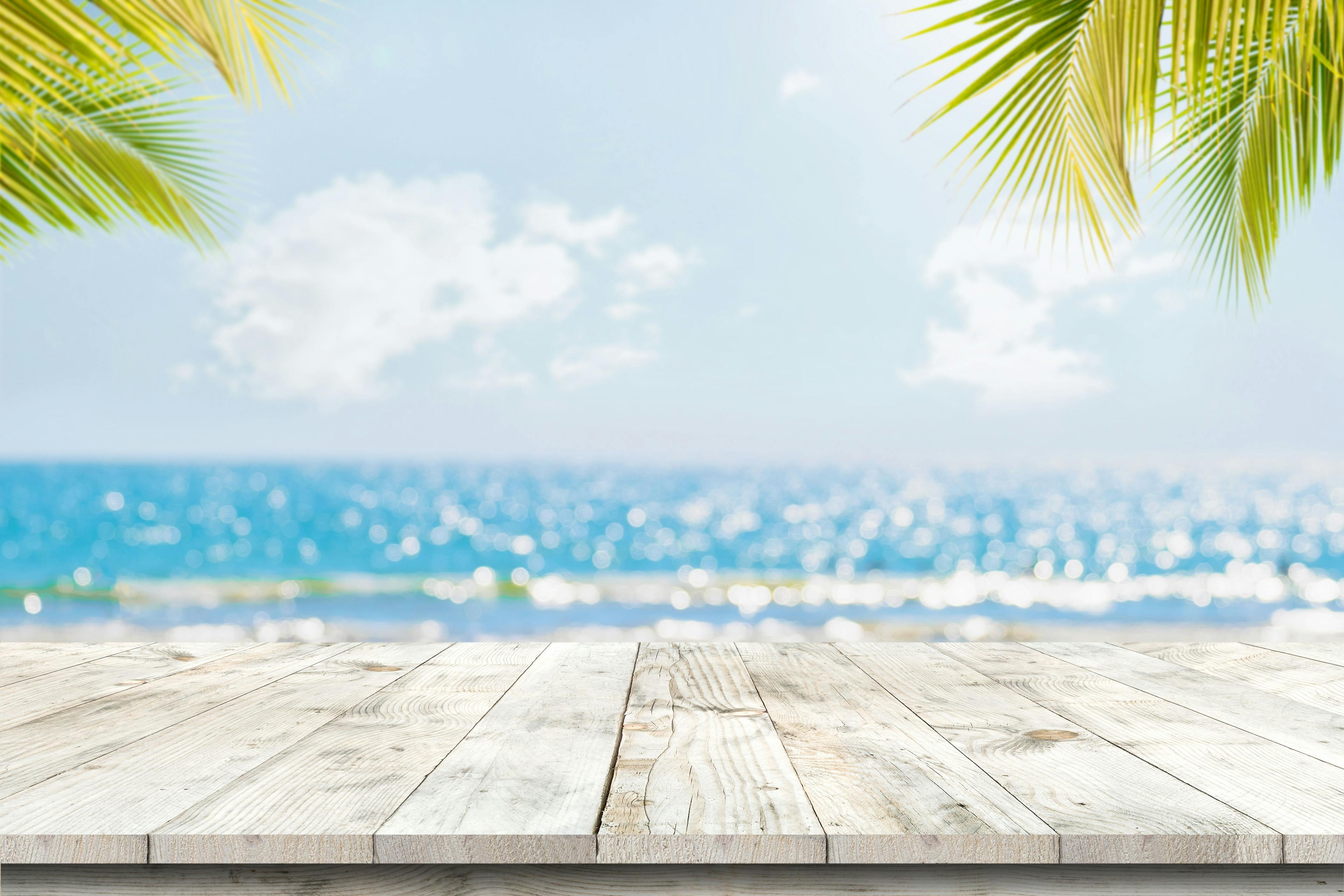 Top of wood table with seascape and palm leaves, blur bokeh light of calm sea and sky at tropical beach background. Empty ready for your product display montage. summer vacation background concept - Image credit: jakkapan | stock.adobe.com