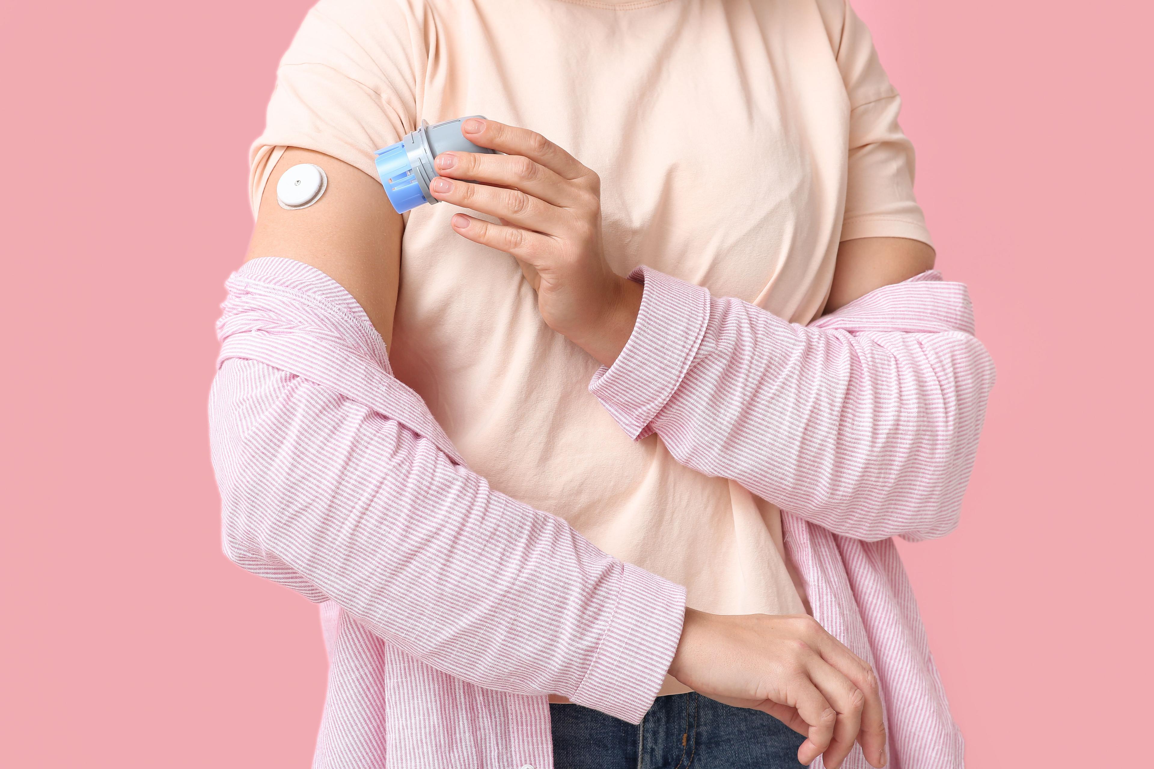 Woman with glucose sensor for measuring blood sugar level and applicator on pink background. Diabetes concept